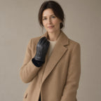 Woman in a camel coat with a gloved fist raised to her collar, black goatskin leather glove with seams, snap strap and fleece cuff